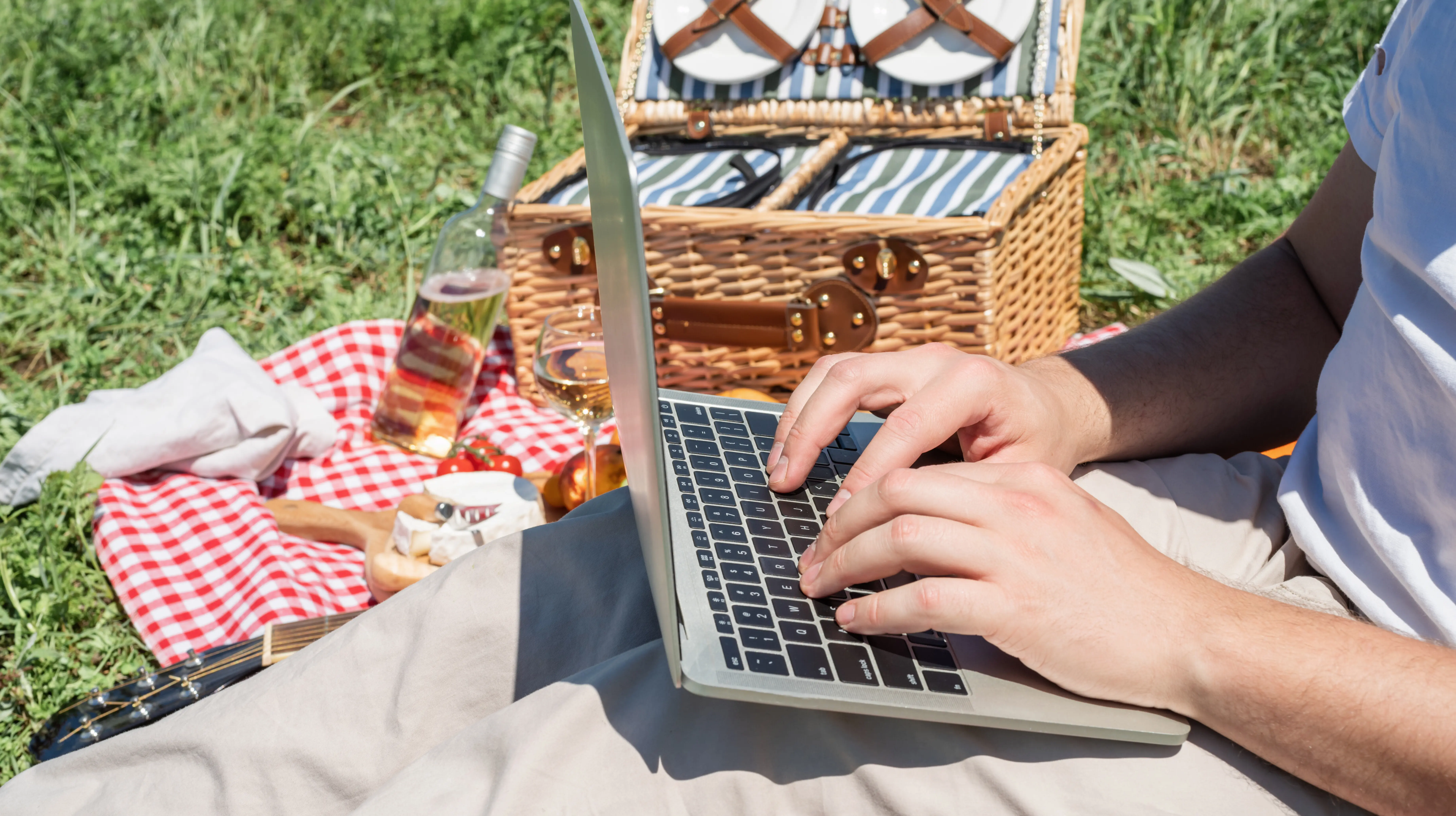 Nicht erkennbarer Mann in weißer Hose bei einem Picknick im Freien, der am Laptop arbeitet.