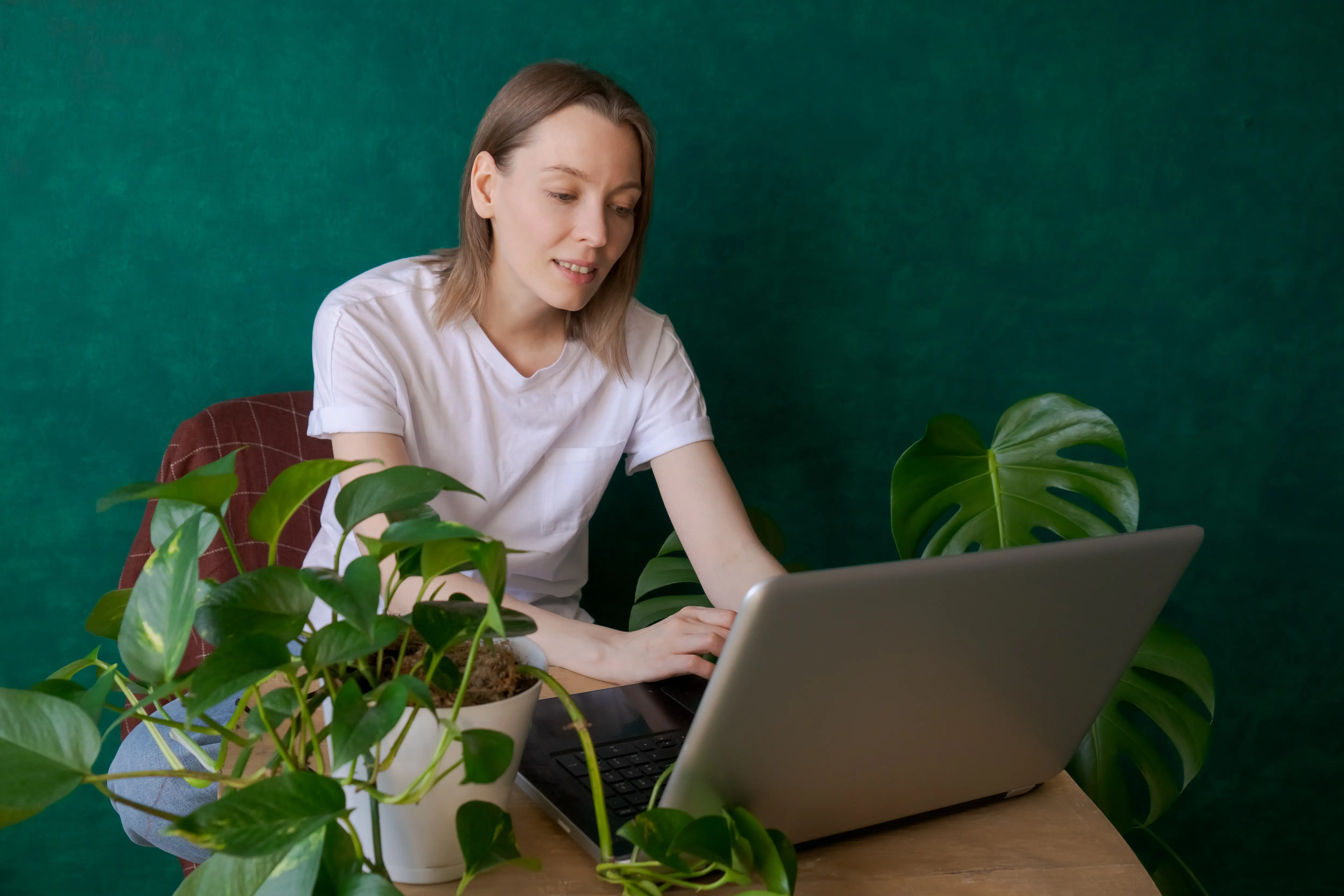 Eine junge Frau sitzt zu Hause am Tisch mit einem Laptop, neben Zimmerpflanzen in Töpfen.