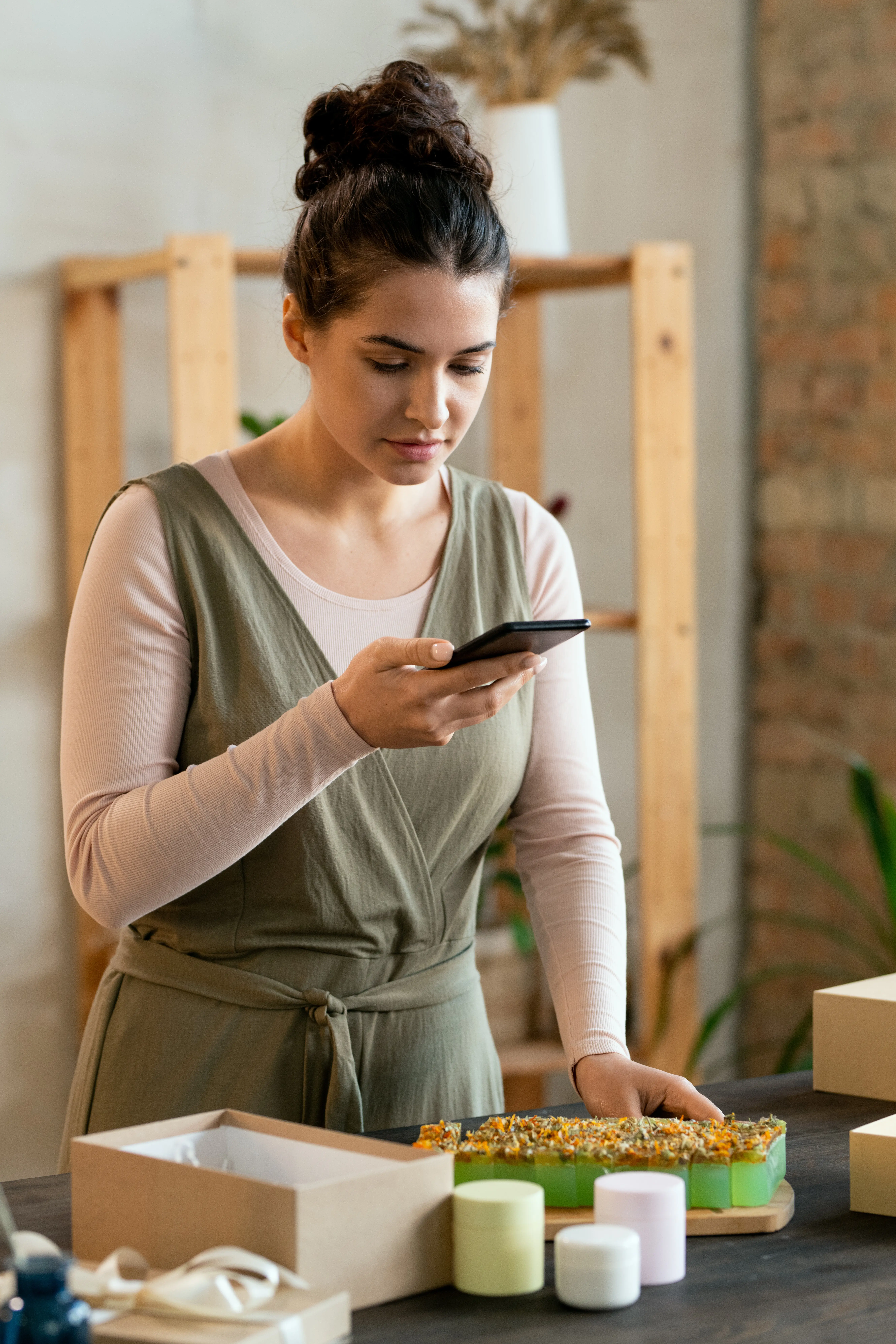 Eine junge Frau steht an einem Tisch mit frischer, aromatischer, handgemachter Seife und Kerzen und filmt mit dem Smartphone, bevor sie diese verpackt.